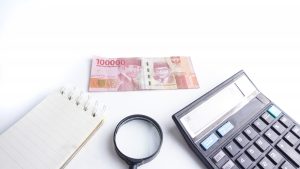 Money concept. Black calculator, Indonesia Rupiah banknote, magnifying glass and notebook on white background