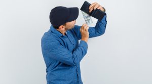 Young man in blue shirt, cap taking dollar bill out of wallet and looking focused , front view.